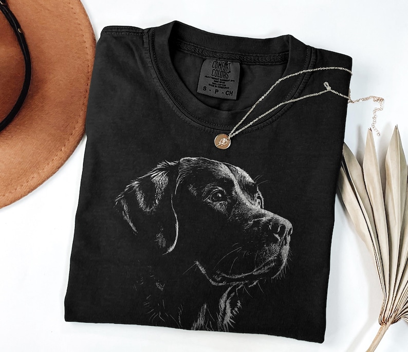Op de afbeelding: Een zwart T-shirt met korte mouwen en een witte lijntekening van een hondenkop op de voorzijde. Het label in de nek bevat de tekst Comfort Colors, S-P-CH en 100% cotton. Het T-shirt is gemaakt van een effen zwarte stof.