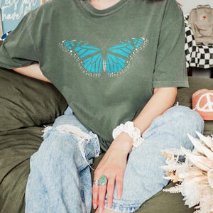 May include: A short-sleeved, crew-neck t-shirt in a muted olive green color. The center of the shirt features a graphic print of a large butterfly with vibrant blue wings, detailed with black veins and white spots along the outer edges of the wings.