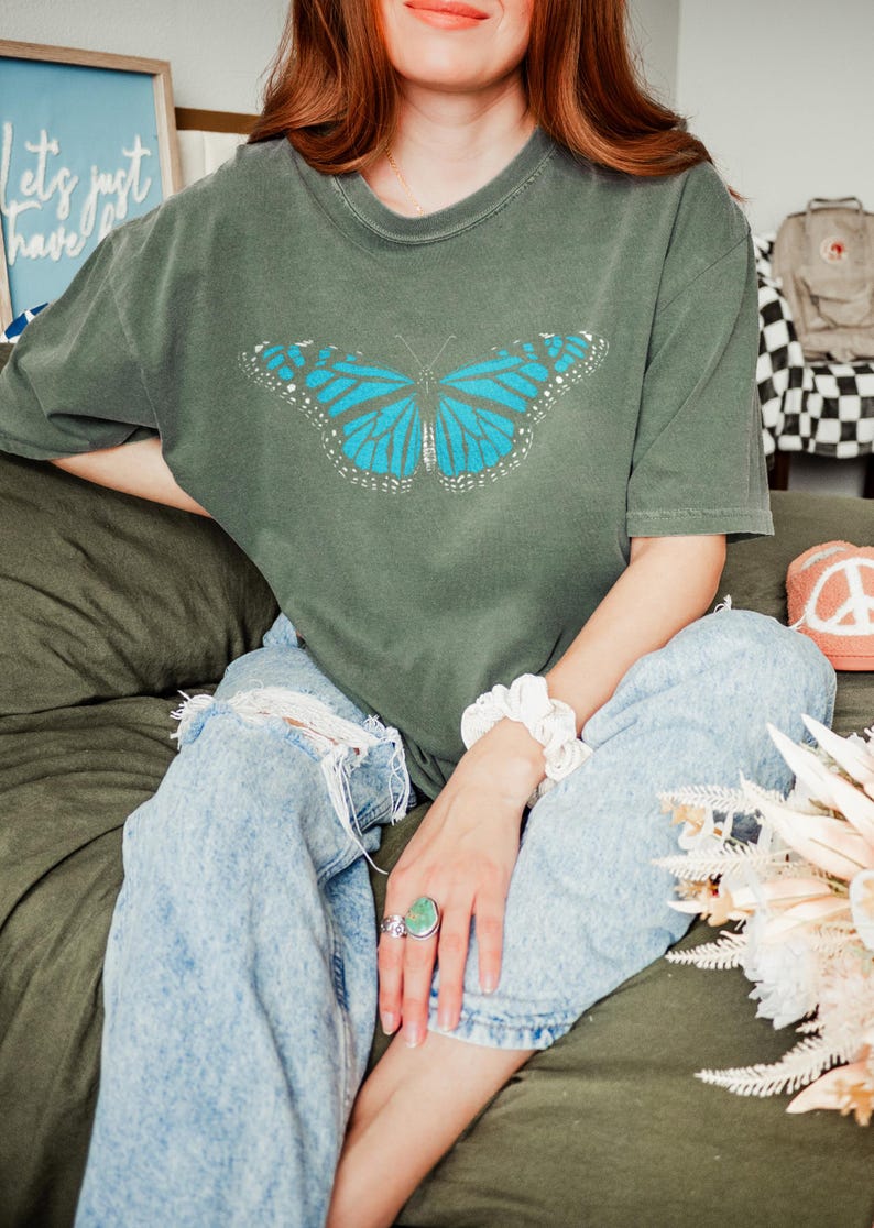 May include: A short-sleeved, crew-neck t-shirt in a muted olive green color. The center of the shirt features a graphic print of a large butterfly with vibrant blue wings, detailed with black veins and white spots along the outer edges of the wings.