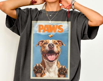 May include: A charcoal gray t-shirt featuring a rectangular graphic print of a brown and white pit bull dog with its paws pressed against a surface. Above the dog, the word PAWS is printed in large orange letters with a small treat icon.