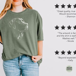 Op de afbeelding: Een olijfgroen T-shirt met korte mouwen en een witte lijntekening van een hondenkop op de borst. Rechts staan drie sets van vijf zwarte sterren met klantbeoordelingen: Great quality, true to size. Would recommend! - Shannon; The artwork is fantastic and the shirt is really soft. Washes well. - Nicole; Beyond expectations. - Kally.
