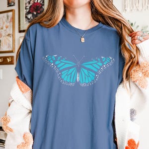 May include: A blue short-sleeve t-shirt featuring a large, centered graphic of a butterfly with bright turquoise wings and white detailing along the edges. The shirt has a crew neckline and a relaxed fit, showcasing a clear, symmetrical insect design.