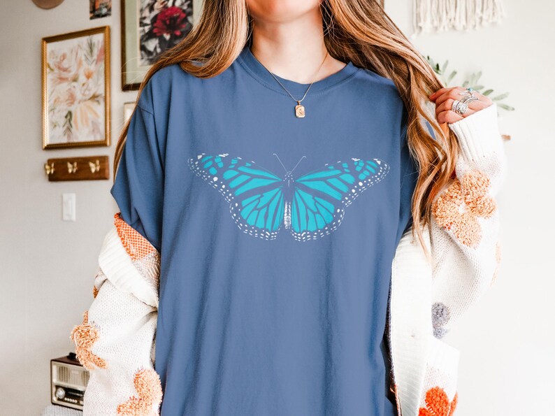May include: A blue short-sleeve t-shirt featuring a large, centered graphic of a butterfly with bright turquoise wings and white detailing along the edges. The shirt has a crew neckline and a relaxed fit, showcasing a clear, symmetrical insect design.