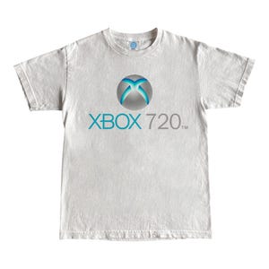 May include: A white t-shirt featuring the Xbox logo in a circle with blue and silver accents, and the text "XBOX 720" in teal and gray. The shirt has a crew neck and short sleeves.