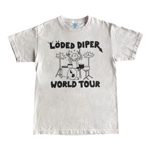 May include: Off-white t-shirt featuring a black graphic of a stick figure playing drums with the text "LÖDED DIPER WORLD TOUR". The shirt has a crew neck and short sleeves. The design includes musical notes and a drum set.