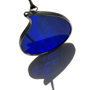 May include: A stained glass pendant with a deep blue hue, suspended by a black cord. The pendant has a unique, curved shape and is framed with a silver-colored metal. The pendant casts a shadow on the white background.