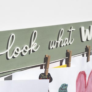 Könnte beinhalten: Ein holzfarbenes Schild in Salbeigrün mit den Worten "look what WE" in weißer Schrift und Blockbuchstaben. Wäscheklammern halten Kunstwerke und Notizen an einer weißen Schnur fest. Die Kunstwerke umfassen ein rosa Herz und andere farbenfrohe Zeichnungen.