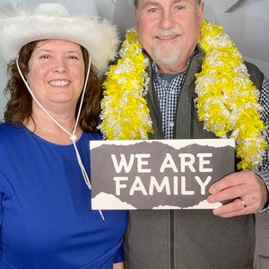 Może przedstawiać: Para w pozie do zdjęcia trzymając tabliczkę z napisem "We Are Family". Kobieta ma na sobie biały kowbojski kapelusz i niebieską sukienkę. Mężczyzna ma na sobie szary kamizelkę i ż&oacute;łto-biały naszyjnik.