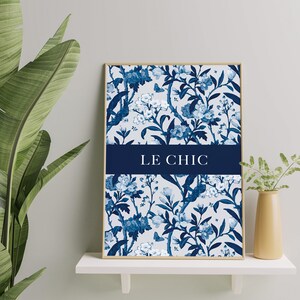 Op de afbeelding: Een blauwe en witte poster met een bloemenprint en de tekst "LE CHIC" in een donkerblauwe banner. De poster is ingelijst in een licht houten lijst en wordt getoond op een witte plank.