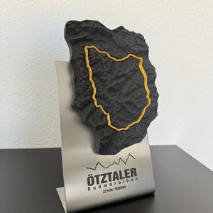 Può includere: Trofeo commemorativo Ötztaler Radmarathon nero e giallo. Il trofeo presenta una mappa in rilievo dettagliata con un contorno del percorso giallo, montato su un supporto in metallo spazzolato. Il supporto mostra il nome e la distanza dell'evento.