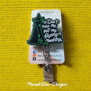May include: A green and black glitter badge reel featuring a silhouette of the Wicked Witch of the West from the Wizard of Oz. The text "Don't make me get my flying monkeys" is written in green glitter.