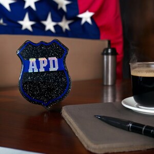 May include: A black and blue police badge with the letters "APD" in white. The badge is sitting on a wooden table with a notebook and pen.