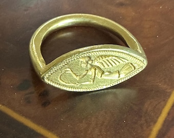 May include: A gold-toned ring featuring an oval-shaped bezel with a textured border. The center of the bezel displays a relief engraving of a winged figure in profile, depicted in a reclining position while holding a curved object in their hands.
