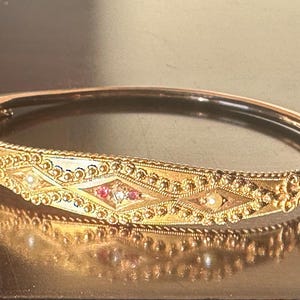 May include: A gold-coloured hinged bangle bracelet with detailed filigree and diamond-shaped patterns. The bracelet features small red and white gemstones, and is a piece of jewellery.