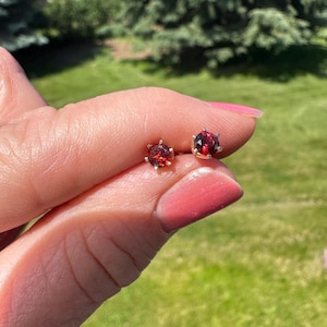Peut inclure: Une paire de petites boucles d'oreilles à clous avec des pierres précieuses grenat rouge foncé, rondes et facettées, serties dans des griffes en métal doré. Chaque boucle d'oreille possède un sertissage classique à six griffes qui maintient la pierre circulaire.