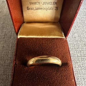 Può includere: Un anello a fascia in oro lucido riposto in un portagioie foderato in velluto marrone scuro. L'interno del coperchio della scatola presenta un testo stampato che recita UHREN-JUWELEN Graz, Jakominiplatz 25, su sfondo in tessuto color crema.
