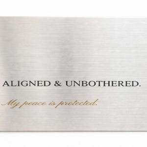 May include: A rectangular, brushed metal card with rounded corners. The text "ALIGNED & UNBOTHERED" is printed in a dark font, with "My peace is protected" in a cursive gold font.