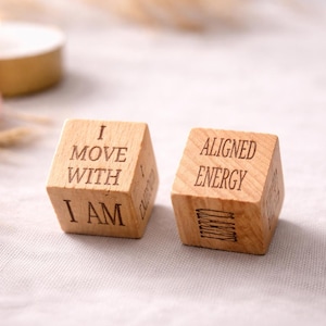 May include: Two wooden dice with engraved text. One die reads "I MOVE WITH I AM", the other says "ALIGNED ENERGY". A lit candle and a pink stone are in the background. The dice are on a white surface.
