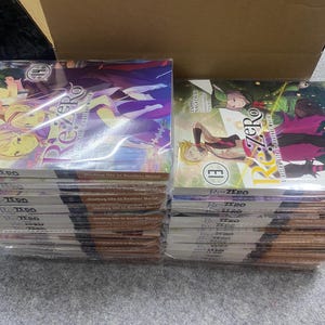 Może przedstawiać: Stosy tomów mangi, z kolorowymi okładkami przedstawiającymi postacie i tytuł "Re:Zero". Książki są zafoliowane i ułożone przed kartonowym pudełkiem. Widoczne są tomy 14 i 13.