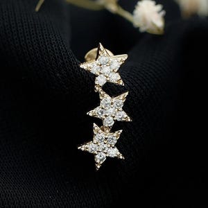 Può includere: Un orecchino a lobo allungato color oro con tre ciondoli a forma di stella collegati, ciascuno incastonato con piccole pietre trasparenti. Il design segue un allineamento verticale, mostrando una finitura metallica e accenti scintillanti.