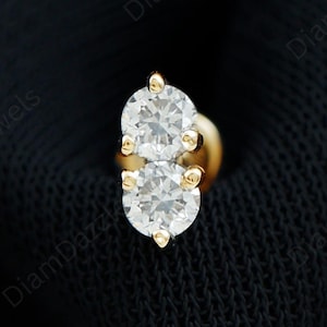 Puede incluir: Un pendiente de nariz dorado con dos gemas redondas y transparentes. El pendiente está engastado en una base dorada y se muestra sobre un fondo oscuro. La joya está diseñada para piercings en la nariz.