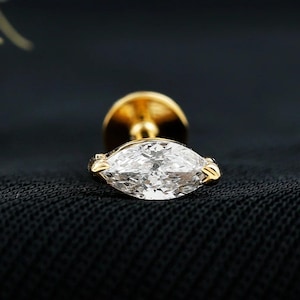 以下が含まれることがあります： 金属製のセッティングにマーキスカットの透明な宝石をあしらったゴールドのスタッドピアス。ジュエリーは白い花を添えた暗い質感の背景に配置されており、DIAM DAZZLE JEWELSというテキストが確認できます。