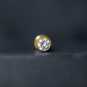 May include: A gold stud earring featuring a single round, clear, faceted gemstone set in a smooth, polished gold bezel setting. The jewelry piece is positioned centrally, showcasing a bright, reflective surface and a classic, circular design.