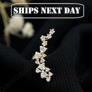 May include: A gold-toned ear climber earring featuring a curved arrangement of multiple small, clear, round-cut stones. The jewelry piece is set against a dark, textured fabric background, showcasing a delicate, branching design with sparkling accents.