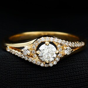 Op de afbeelding: Een gouden ring met een ronde diamant in het midden, omringd door kleinere diamanten. De ring heeft een gespleten band met een diamant accent aan elke kant van het middelste stuk.