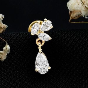 May include: A gold-tone earring featuring a cluster of three small marquise-cut clear stones at the top, with a single dangling pear-cut clear stone suspended below. The piece is set in a metallic gold-tone frame and includes a flat circular backing.