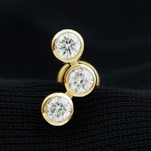 May include: A gold earring featuring three round, bezel-set clear stones arranged in a curved, vertical cluster. The stones are set in polished yellow gold, creating a bright, reflective surface. The text DIAM DAZZLE JEWELS appears in the corner.