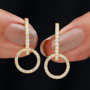 Puede incluir: Un par de pendientes de aro dorados, cada uno con una barra vertical y un aro circular, ambos engastados con pequeñas piedras preciosas transparentes. Los pendientes se muestran sobre un fondo negro.