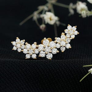 Könnte beinhalten: Ein goldfarbener Ohrschmuck mit floralem Design, besetzt mit zahlreichen kleinen, funkelnden weißen Edelsteinen. Das Schmuckstück ist vor einem schwarzen Hintergrund mit weißen Blumen im Hintergrund platziert.