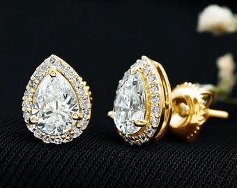 May include: A pair of gold stud earrings with pear-shaped, clear gemstones. Each earring is surrounded by a halo of smaller, clear stones. The earrings are set against a black background, with white flowers in the background.