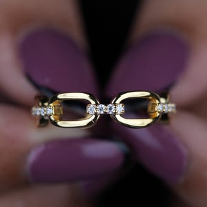 May include: A gold-colored ring with a chain link design, adorned with small, sparkling gemstones. The ring is held between two hands with purple nail polish, creating a contrast with the jewelry.