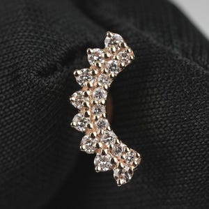 Può includere: Un orecchino curvo color oro con una serie di piccole pietre preziose trasparenti. Le pietre preziose sono disposte in un motivo graduato, creando un design scintillante ed elegante. L'orecchino è mostrato su uno sfondo scuro.