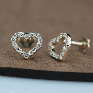 以下が含まれることがあります： 金色のハート型のスタッドピアス。各イヤリングは、小さな透明な宝石で縁取られています。イヤリングは茶色の表面に表示されています。