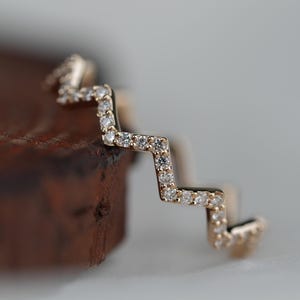 Può includere: Un anello color oro con un design a zig-zag, incastonato con piccole pietre chiare e scintillanti. Il design dell'anello è evidenziato su uno sfondo sfocato, sottolineando i dettagli intricati. L'anello sembra essere in oro ed è un gioiello raffinato.