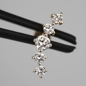 May include: A curved earring featuring five round-cut clear stones set in a gold-toned metal bar. The stones are arranged in a graduated size pattern, with the largest stone positioned near the center and smaller stones tapering toward both ends.