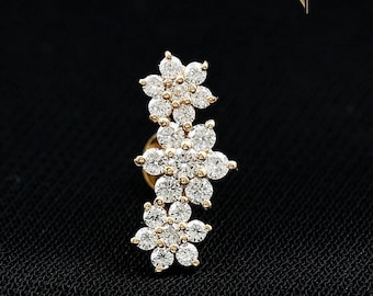 May include: A gold earring stud featuring three connected flower shapes, each set with multiple round clear stones. The design is vertical and includes the text DIAM DAZZLE JEWELS in the upper right corner. The piece has a metallic finish.