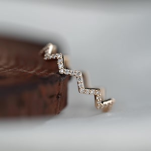 Puede incluir: Un anillo dorado con un diseño en zigzag, incrustado con pequeños diamantes brillantes. El diseño del anillo se destaca sobre un fondo borroso. Una joya elegante con detalles delicados.