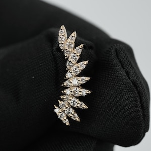 Op de afbeelding: Een gouden ear climber-oorbel met een gebogen, waaiervormige opstelling van meerdere heldere stenen in marquise-slijpvorm. Het sieraad is ontworpen om de vorm van het oor te volgen, waarbij elke steen in een gepolijste gouden zetting is geplaatst.