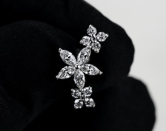 May include: A silver-toned earring featuring a central flower design composed of five marquise-cut clear stones, flanked by two smaller four-petaled flower motifs made of round clear stones. The piece has a metallic finish and clear faceted stones.