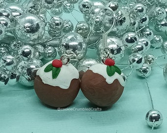 May include: A pair of Christmas pudding earrings. Each earring is a miniature, round, brown pudding with white icing, a red cherry, and green holly leaves. The earrings are set against a backdrop of silver ornaments.
