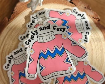 以下が含まれることがあります： 「Comfy and Cozy」という文字が書かれた、ピンクとブルーのセーターの形をしたステッカーが4枚セットになっています。ステッカーは木製の表面に置かれています。