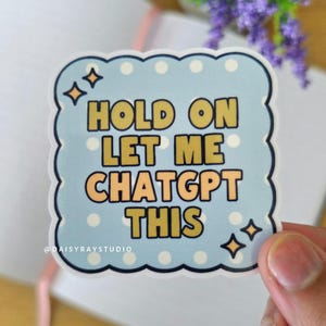 May include: A light blue sticker with a scalloped edge and the text "HOLD ON LET ME CHATGPT THIS" in yellow and orange. The sticker has white polka dots and gold star accents. The sticker is held by a hand.