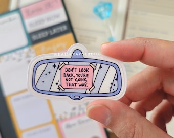 May include: A clear sticker in the shape of a car mirror with a pale blue border. The sticker features a pink rectangle with the text "DON'T LOOK BACK, YOU'RE NOT GOING THAT WAY." The sticker is held in front of a blurred background.