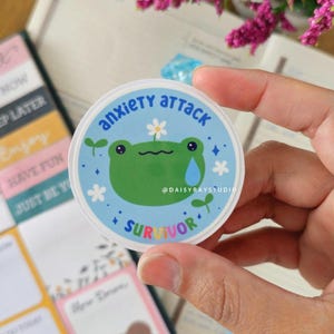 May include: A circular sticker with the words "anxiety attack" and "survivor". The design features a green frog with a tear and a daisy, set against a blue background. The sticker is held in a hand.