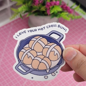 Peut inclure: Un autocollant représentant une illustration de dessins animés de petits pains chauds dans une poêle bleue. Les petits pains sont brun clair avec des croix blanches. Le texte "I LOVE YOUR HOT CROSS BUNS" est imprimé au-dessus de la poêle en bleu.