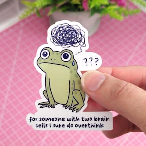 May include: A die-cut vinyl sticker featuring a light green frog with a confused expression, a blue sweat drop, a dark blue scribble representing a tangled thought, three question marks, and the text: for someone with two brain cells I sure do overthink.